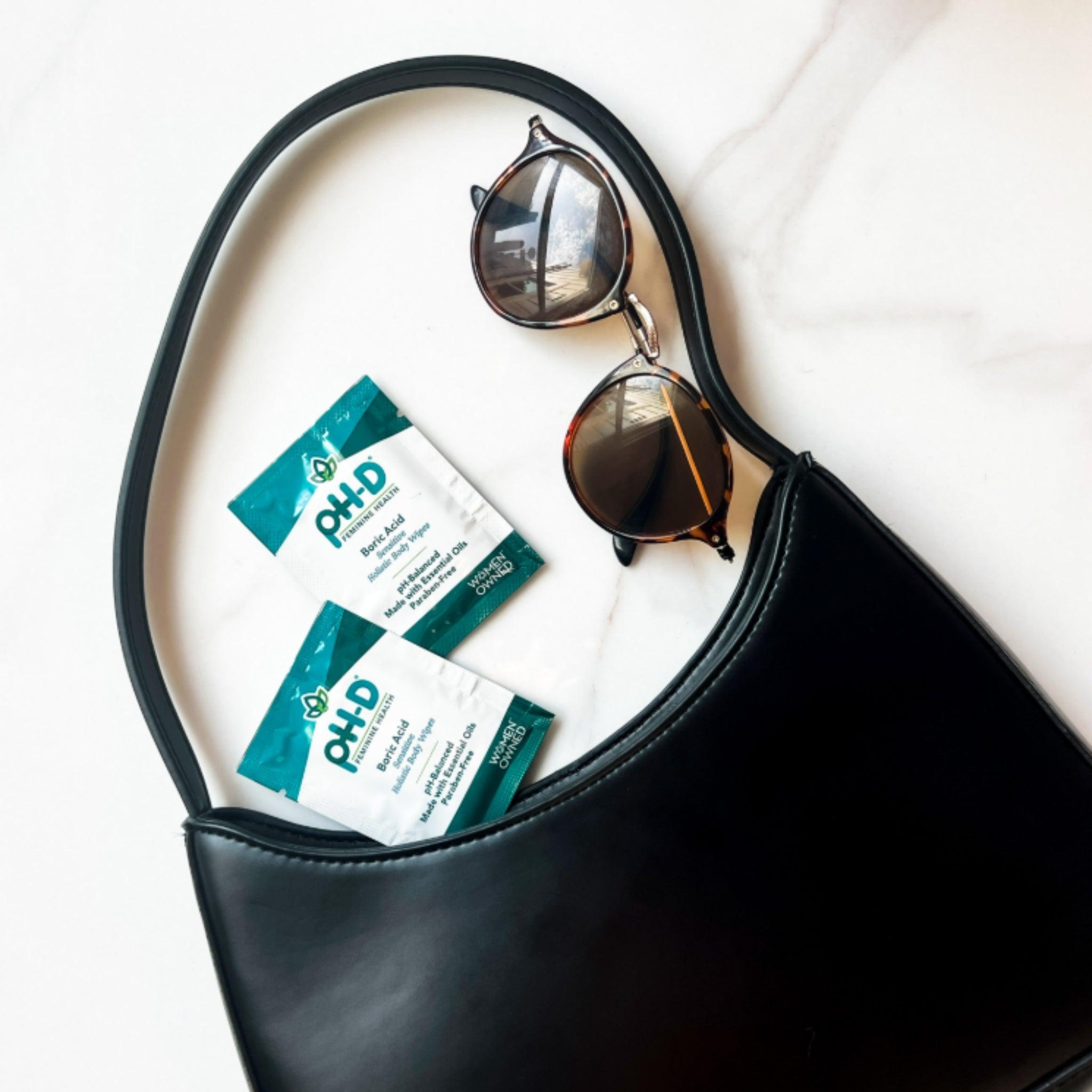 pH-D Boric Acid Holistic Body Wipes packets displayed with a stylish black handbag and sunglasses, emphasizing portability and on-the-go feminine care.