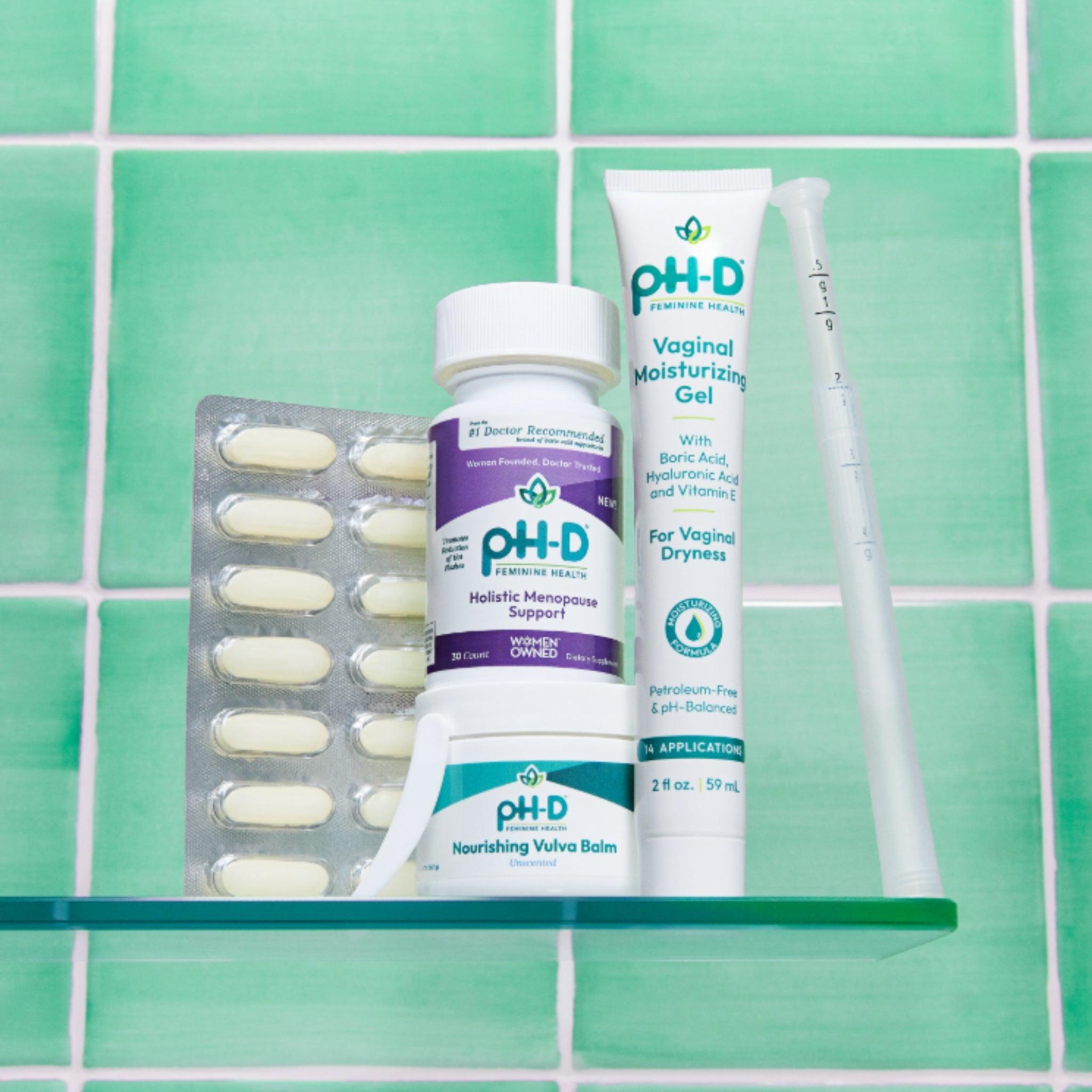 A bathroom shelf displaying a selection of pH-D Feminine Health products: Holistic Menopause Support capsules, a tube of Vaginal Moisturizing Gel, Nourishing Vulva Balm, and a blister pack of supplements. The green tiled background creates a clean, refreshing aesthetic, emphasizing the products' health and wellness focus.